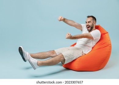 Full Length Young Smiling Happy Man 20s In White T-shirt Sit In Bag Chair Play Racing Game Hold Virtual Steering Wheel Rest Relax Isolated On Plain Pastel Light Blue Color Background Studio Portrait.