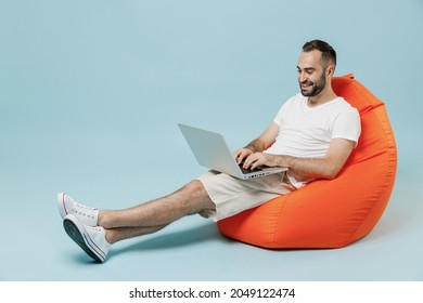 Full Length Young Smiling Happy Man 20s In Casual White T-shirt Sit In Bag Chair Hold Use Work On Laptop Pc Computer Rest Relax Isolated On Plain Pastel Light Blue Color Background Studio Portrait.