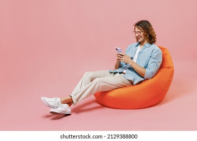 Full Length Young Smiling Happy Man With Long Curly Hair Wear Blue Shirt White T-shirt Glasses Sit In Bag Chair Hold In Hand Use Mobile Cell Phone Isolated On Pastel Plain Pink Color Background Studio