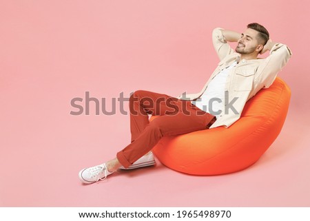 Full length young smiling happy overjoyed joyful fashionable man 20s in jacket white t-shirt sitting in bean bag chair resting hold hand behind neck isolated on pastel pink background studio portrait.