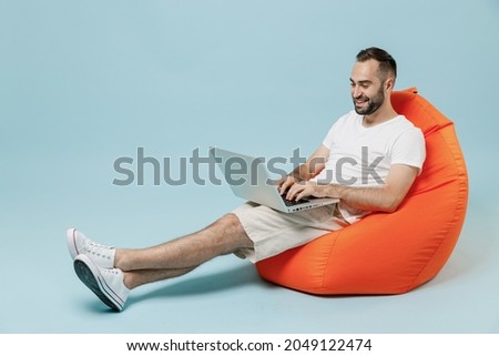 Full length young smiling happy man 20s in casual white t-shirt sit in bag chair hold use work on laptop pc computer rest relax isolated on plain pastel light blue color background studio portrait.