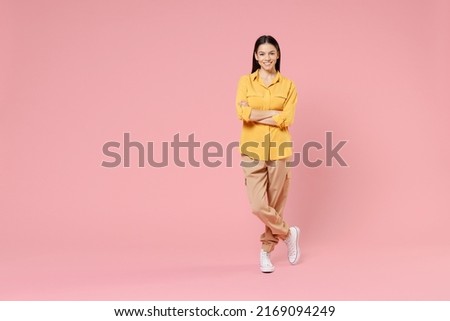 Full length of young smiling pretty happy brunette positive attractive beautiful latin woman 20s in yellow casual shirt hold hands crossed folded isolated on pastel pink background studio portrait