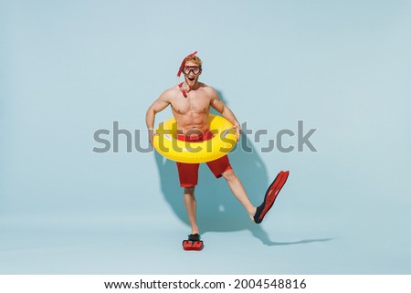 Full length young sporty man in red shorts swimsuit diving mask relax near pool hold inflatable ring flippers raise up leg isolated on pastel blue background. Summer vacation sea rest sun tan concept.