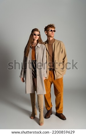 full length of young and stylish couple in autumnal outfits posing on grey