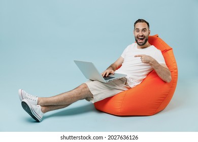Full Length Young Surprised Happy Man In Casual White T-shirt Sit In Bag Chair Hold Use Work Point Index Finger On Laptop Pc Computer Rest Relax Isolated On Plain Pastel Light Blue Background Studio.