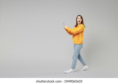 Full length of young surprised shocked caucasian happy woman 20s wear knitted yellow sweater going walking going hold mobile cell phone look camera isolated on grey color background studio portrait.