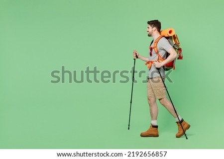 Full length young traveler white man carry backpack stuff mat walk with trakking poles isolated on plain green background. Tourist leads active healthy lifestyle. Hiking trek rest travel trip concept