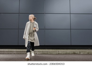 full length of young woman with blonde hair with bangs walking in coat, black leather pants, hoodie and boots while holding paper cup with takeaway coffee near grey modern building on street - Powered by Shutterstock