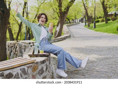 Full Length Young Woman In Green Jacket Jeans Sit On Bench In City Spring Park Outdoors Resting Eat Fast Food Hamburger Have Breakfast Waving Hand Say Hello People Unhealthy Urban Lifestyle Concept.