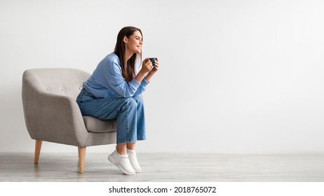 Full length of young woman having coffee break in cozy armchair against white studio wall, banner design with free space. Charming millennial lady relaxing, enjoying hot drink - Shutterstock ID 2018765072
