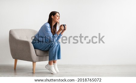 Full length of young woman having coffee break in cozy armchair against white studio wall, banner design with free space. Charming millennial lady relaxing, enjoying hot drink