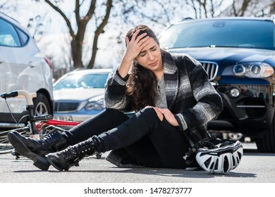 Full length of a young woman sitting on the asphalt with severe headache and dizziness after bicycle accident on a busy city street