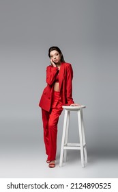 full length of young woman in trendy red suit posing near white chair on dark grey