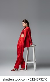 full length of young woman in trendy red suit standing near white chair on dark grey