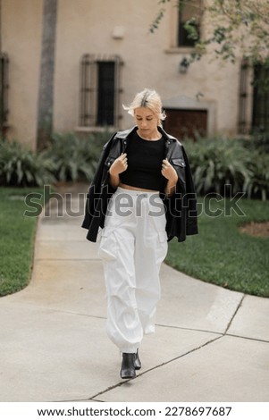 full length of young woman in white cargo pants and leather shirt jacket walking near house in Miami