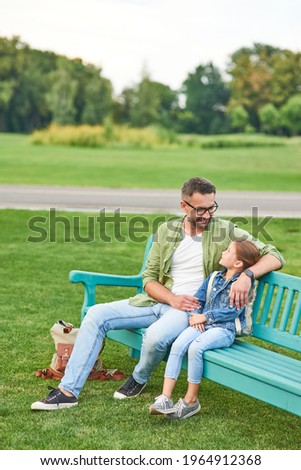 Full lentgh shot of happy young father and his cute little daughter talking while sitting on the bench, spending time together in the green park