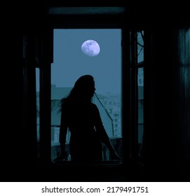 Full Moon In The Night Dark  Blue  Sky. Black Silhouette Of Young Slim Magical Whitch Woman Looking Out Of An Open Balcony Window. City, Town With Many Houses On Background.