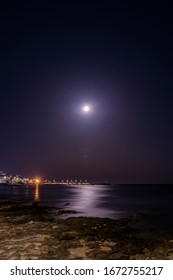 The Full Moon Is Reflected On The Water Of The Sea, In A Beautiful Night Landscape. In Italy, Puglia, Lecce, Salento. The Marina With Boats Moored In The Background. The Rocks In The Foreground.