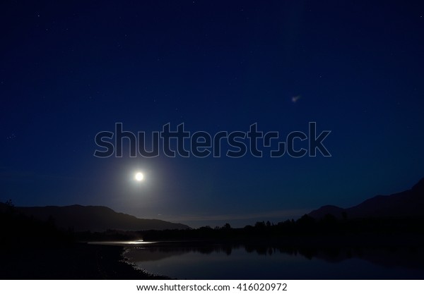 full moon reflection in river surface at night\
surrounded by stars and\
mountains
