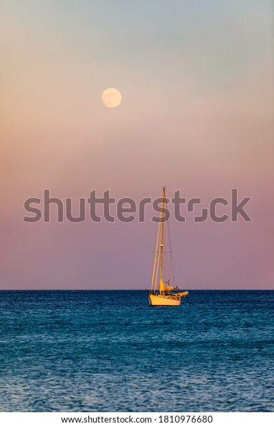 Full moon rising over the water with a small\
sailing boat in the foreground. Sailing boat with raising moon at\
sunset. Moon rising over the sea and yacht floating on the water\
surface. Sardinia, Italy
