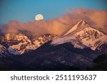 Full moon setting into the early morning colorful sunrise alpenglow on the clouds and snow covered Rocky Mountain peaks 