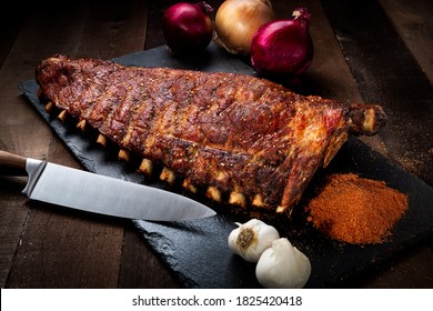 Full Rack Of Pork Ribs, Styled On A Black Slate With Garnish And Utensil.