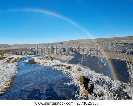 A full rainbow arches over the dramatic black basalt canyon of Dettifoss waterfall in northern Iceland, with white frost crystals covering the surrounding vegetation and a vivid blue glacial river below.