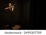 Full shot of concentrated female violinist in elegant dress playing classical music standing alone under projector light in dark studio with black curtains, copy space