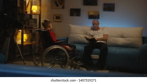 Full Shot Of An Elderly Man Reading A Book To A Disabled Boy On A Wheelchair During A Storm