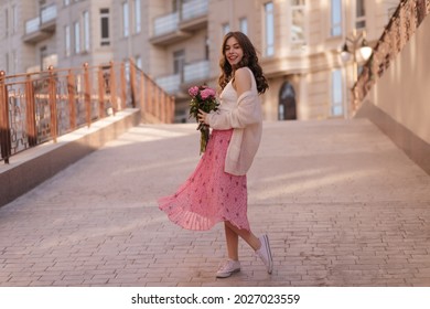 Full shot portrait of young surprised teen girl holds a peonies bouquet in her hands. Impressively she stands in a half turn, dressed in pretty pinky skirt, white undershirt and purse beige sweater 
