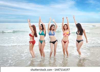 full shot of young asian beautiful and joyful girls in bikini who dancing and raising hands on tropical beach with happy and smile faces on holiday vacation with blue sky(party and friendship concept)