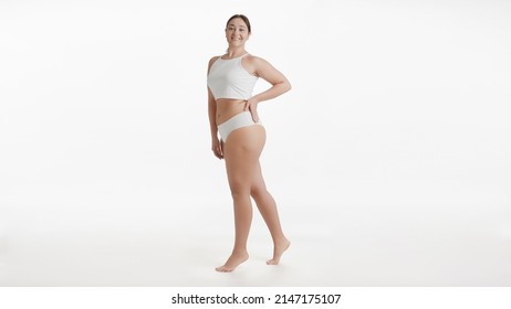 Full shot of young pretty Caucasian plus size woman in white underwear stands on tiptoes putting hand on her waist smiling wide to the camera on white background | Body care concept