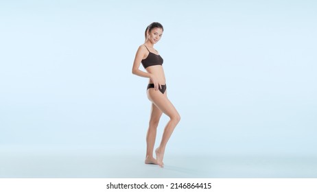 Full shot of young slim good-looking Asian woman in black underwear posing and touching her hip on pale blue background | Leg and body care concept