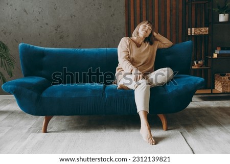 Full size body calm elderly woman 50s years old wears casual clothes sits on blue sofa prop up head close eyes dream stay at home flat rest relax spend free spare time in living room indoor grey wall