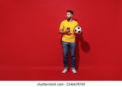 Full Size Body Length Amazed Shocked Surprised Young Bearded Man Football Fan In Yellow T-shirt Look Aside Hold Soccer Ball Use Mobile Cell Phone Isolated On Plain Dark Red Background Studio Portrait