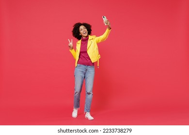 Full size body length bright young curly black latin woman 20s wears yellow jacket doing selfie shot on mobile cell phone post photo on social network isolated on plain red background studio portrait