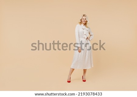 Full size body length charming elderly gray-haired blonde woman lady 40s years old wears pink dress hold hand on waist looking camera posing isolated on plain pastel beige background studio portrait