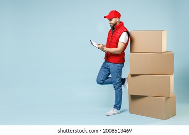 Full Size Body Length Delivery Guy Employee Man In Red Cap White T-shirt Vest Uniform Work As Dealer Courier Hold Blank Cardboard Box Isolated On Pastel Blue Color Background Studio. Service Concept