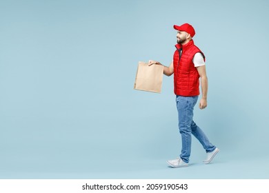 Full size body length delivery man in red cap white T-shirt vest work as dealer courier service hold brown clear blank craft paper takeaway bag mock up isolated on pastel blue color background studio