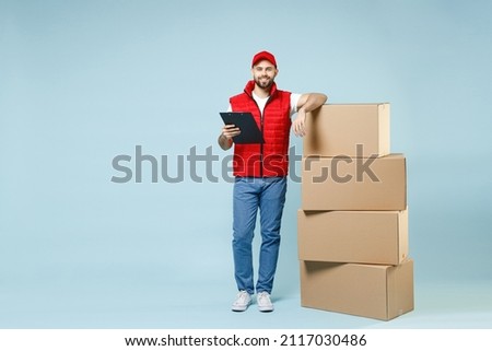 Full size body length delivery guy employee man in red cap white T-shirt vest uniform work as dealer courier hold blank cardboard box isolated on pastel blue color background studio. Service concept