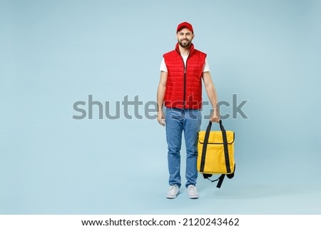 Full size body length delivery guy employee man in red cap white T-shirt uniform work as courier hold yellow thermal food bag backpack isolated on pastel blue color background studio. Service concept