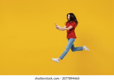 Full Size Body Length Excited Young Brunette Woman 20s Wears Basic Red T-shirt Jump Hold In Hand Use Mobile Cell Phone Isolated On Yellow Background Studio Portrait. People Emotions Lifestyle Concept