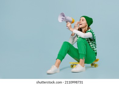 Full size body length excited young brunette girl teen student wears checkered green vest hat sit on skateboard hold scream in megaphone isolated on plain pastel light blue background studio portrait