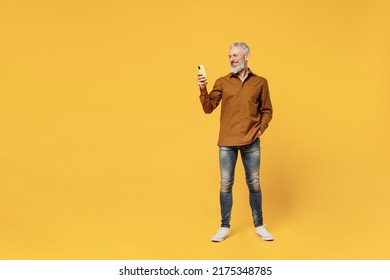 Full size body length excited confident smiling elderly gray-haired bearded man 50s years old wears brown shirt hold in hand use mobile cell phone isolated on plain yellow background studio portrait
