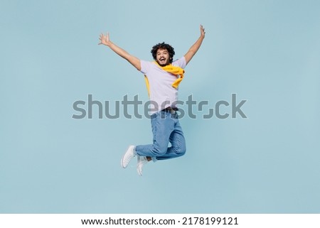 Full size body length exultant cheerful young bearded Indian man 20s years old wears white t-shirt raise hands palms up looking camera isolated on plain pastel light blue background studio portrait