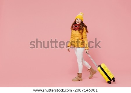 Full size body length fascinating young woman 20s years old wears yellow jacket hat mittens hold push suitcase bag walking stepping pace isolated on plain pastel light pink background studio portrait