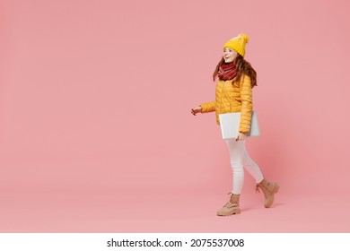 Full size body length fun young woman 20s years old wears yellow jacket hat mittens hold use work on laptop pc computer walking stepping isolated on plain pastel light pink background studio portrait