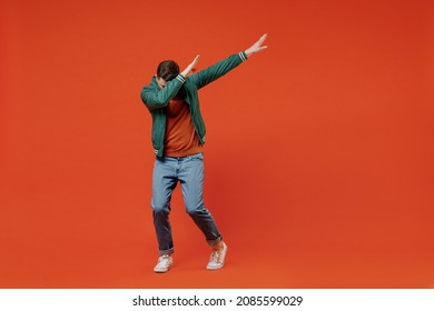 Full size body length fun young brunet man 20s wears red t-shirt green jacket doing dab hip hop dance hands move gesture youth sign hide cover face isolated on plain orange background studio portrait.