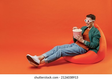 Full Size Body Length Fun Young Brunet Man 20s Wears Red T-shirt Green Jacket Sit In Bag Chairin 3d Glasses Watch Movie Film Hold Bucket Of Popcorn Isolated On Plain Orange Background Studio Portrait.