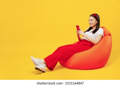 Full Size Body Length Fun Young Girl Woman Of Asian Ethnicity 20s Years Old Wears Casual Clothes Sit In Bag Chair Hold In Hand Use Mobile Cell Phone Isolated On Plain Yellow Background Studio Portrait